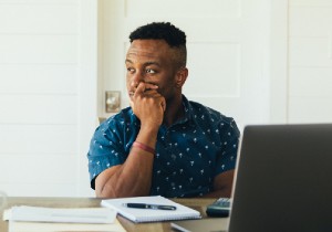 A man sitting down at his desk with his hand over his mouth gazing out in the distance thinking.